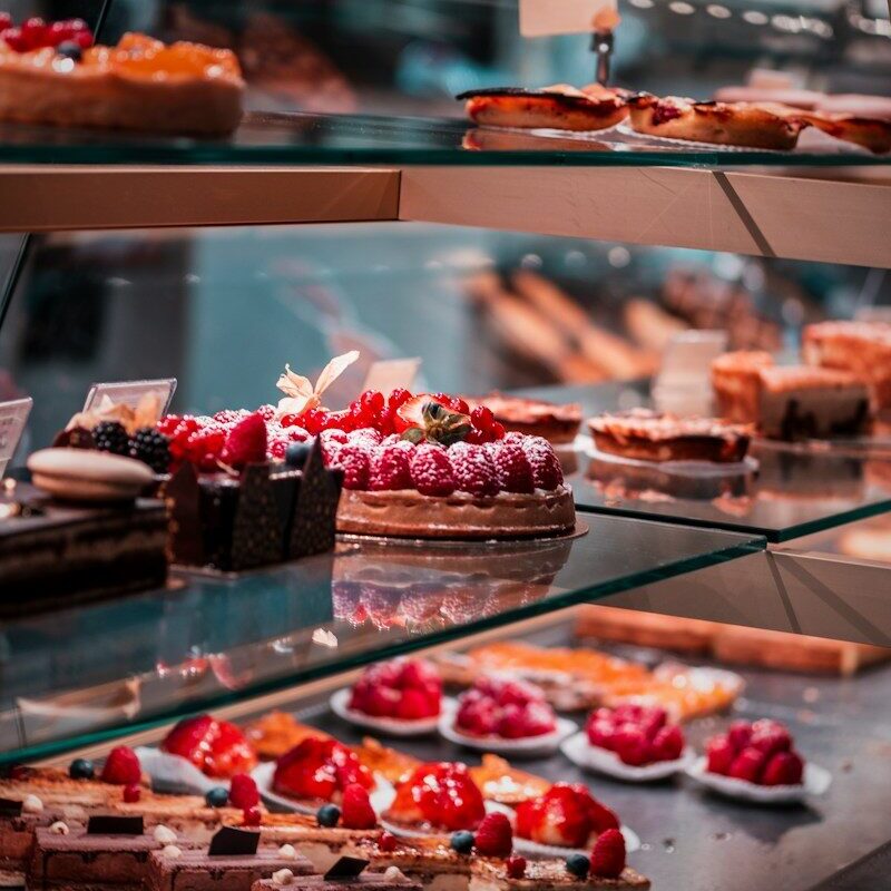 Assorted fruit tarts and chocolate cakes displayed on glass shelves in a bakery